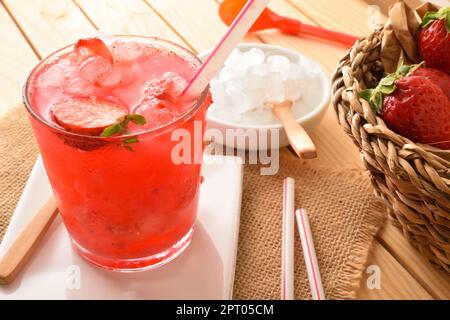 Bicchiere di succo naturale di fragola maturo appena raccolto con ghiaccio su tavolo di legno e contenitore con ghiaccio tritato. Vista in alto. Composizione orizzontale. Foto Stock