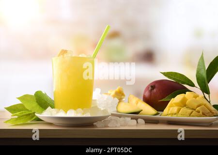 Particolare della bevanda analcolica di mango con molto ghiaccio su una panca di legno della cucina con la frutta ed una ciotola con il ghiaccio intorno esso. Vista frontale. Composizione orizzontale. Foto Stock