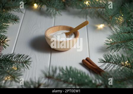 Zucchero in una ciotola di legno con un cucchiaio e un Bastone di cannella accanto ai rami verdi di un albero di Natale Con lampadine a LED su un bac di legno bianco Foto Stock