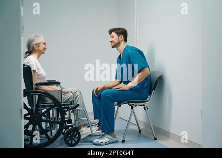 Infermiere in uniforme medica comunica con il paziente anziano in sedia a rotelle Foto Stock