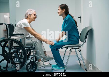 Donna dai capelli grigi che si trova in una sedia a rotelle in clinica Foto Stock