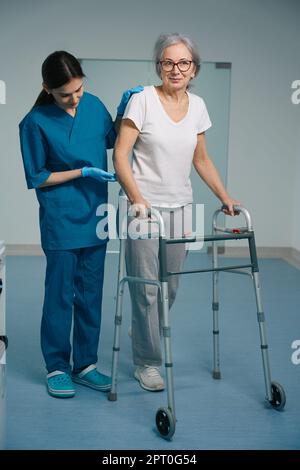 Il paziente anziano cammina con un camminatore lungo il corridoio dell'ospedale Foto Stock