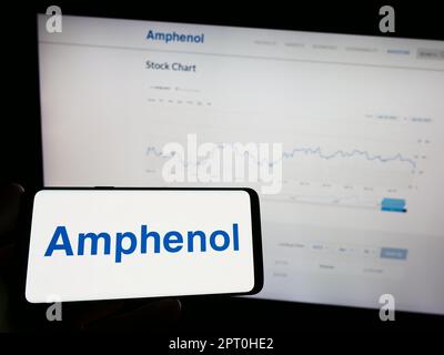 Persona che tiene il telefono cellulare con il logo della società americana di elettronica Amphenol Corporation sullo schermo di fronte alla pagina web. Messa a fuoco sul display del telefono. Foto Stock