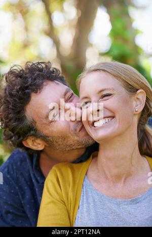 Dandole un grande bacio sulla guancia, un marito affettuoso e una moglie in piedi all'aperto Foto Stock