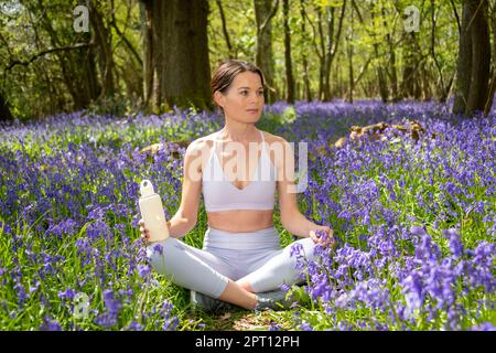 Adatta donna sportiva che beve da una bottiglia d'acqua di vetro, seduto a croce in un campo di bluebells. Foto Stock