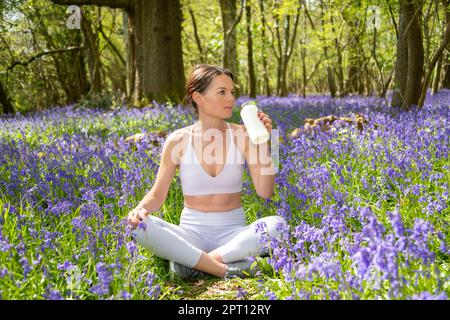 Adatta donna sportiva che beve da una bottiglia d'acqua di vetro, seduto a croce in un campo di bluebells. Foto Stock