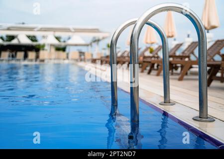 vista prospettica primo piano delle barre di appoggio all'ingresso della piscina all'aperto con acqua blu e terrazza con sedie a sdraio sullo sfondo Foto Stock