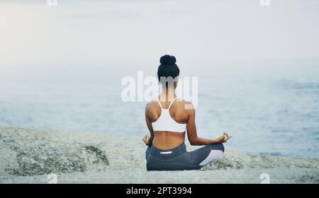 Essere fermi. Ripresa retroversa di una donna irriconoscibile seduta a croce e meditando da sola dall'oceano durante una giornata colta Foto Stock