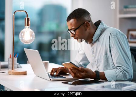 HES è connesso in tutto il mondo aziendale. un bel giovane uomo d'affari che usa il suo portatile e il cellulare mentre lavora tardi nel suo ufficio Foto Stock