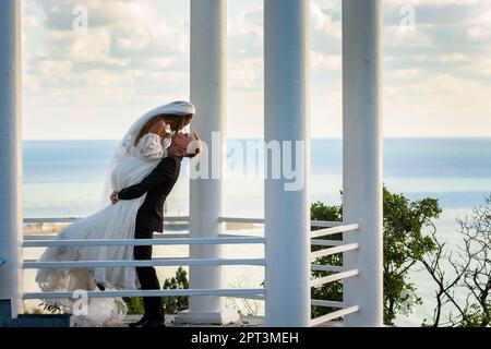 Gli sposi novelli baciano in un bellissimo e pittoresco gazebo sullo sfondo del mare Foto Stock