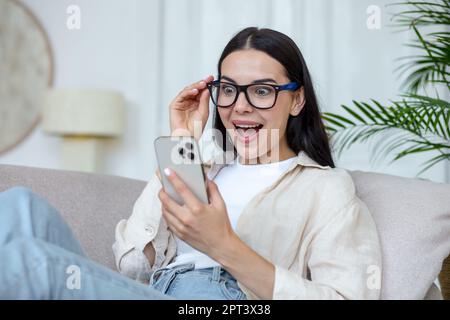 Una giovane donna piacevolmente sorpresa è seduta sul divano a casa e guarda il telefono con gioia. Ricevuto denaro, leggere buone notizie, messaggi. Foto Stock