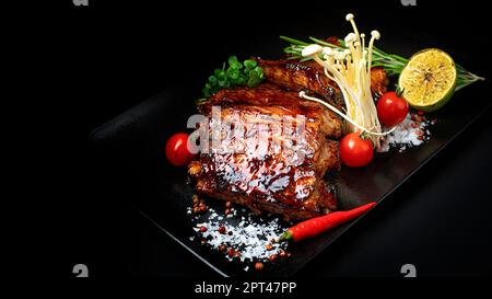 costolette di maiale appetitose con pomodori ciliegini, lime, funghi ed erbe su sfondo nero Foto Stock