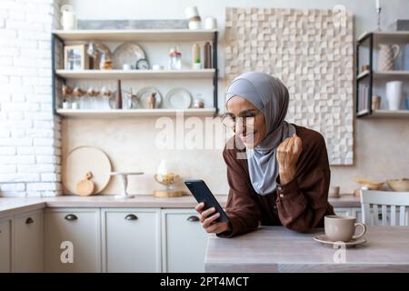 Piacevolmente sorpresa giovane donna musulmana in hijab a casa in cucina guardando il telefono con ammirazione. Ricevuto denaro, leggere buone notizie, messaggi. Mostra un gesto di vittoria con la mano. Foto Stock