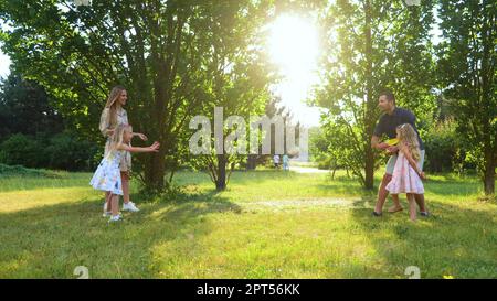 Famiglia positiva lancio disco frisbee nel parco. Ragazza sorridente cattura piastra di frisbee. Giovani genitori che giocano a frisbee con figlie. Divertimento per tutta la famiglia Foto Stock