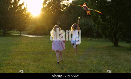 Vista posteriore di bambini felici che corrono sulla natura volando aquilone colorato. Il retro di due simpatiche ragazze gioiosa corrono all'aperto nel parco. Bambini spensierati che giocano avendo Foto Stock