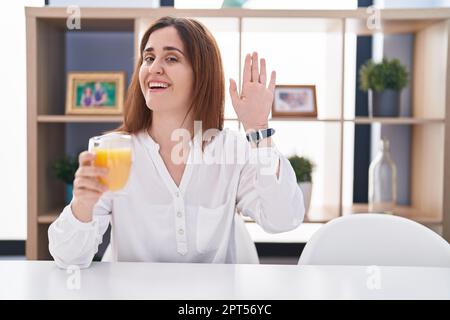 Donna brunette bere un bicchiere di succo d'arancia rinuncia dicendo ciao felice e sorridente, amichevole gesto di benvenuto Foto Stock