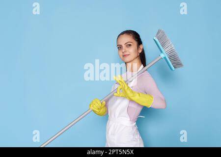 donna caucasica in guanti di gomma e grembiule più pulito che tiene la scopa e mostra ok gestureon sfondo blu. Foto Stock