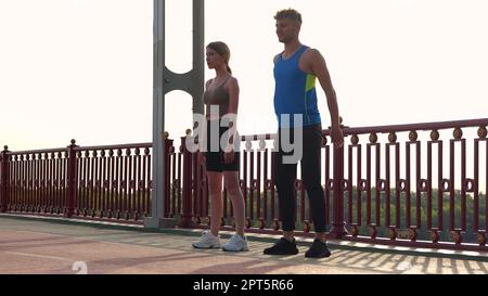 Macchina fotografica avvicinandosi giovane coppia di uomo e donna in abbigliamento sportivo allenarsi fuori sul ponte. Maschio e femmina caucasici sportivi che fanno esercizio fisico Foto Stock