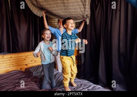 Famiglia felice madre multietnica, padre e bambini, figlio e figlia, ridendo, giocando, combatte i cuscini e salta a letto in camera da letto a casa nel Foto Stock