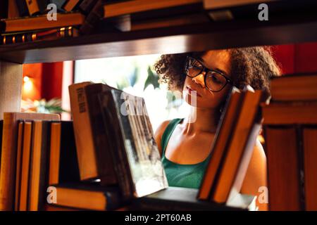 Felice studentessa afro-americana studia in biblioteca universitaria e cerca il libro, indossando gli occhiali Foto Stock