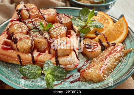 Deliziosi waffle con banana caramellata, arancia tagliata a fette, menta, marmellata e salsa dolce sul piatto su sfondo di legno chiaro. Impostazione della tabella. Dessert. Servizio Foto Stock