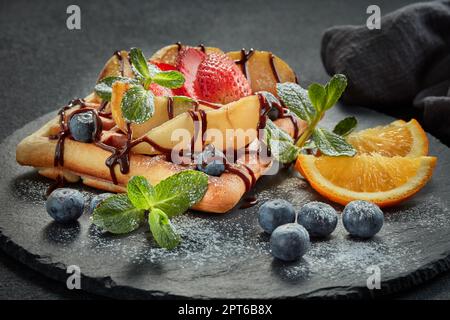 Deliziosi waffle con pera caramellata, frutti di bosco, menta, zucchero a velo e salsa dolce sul piano di servizio nero su sfondo grigio. Dessert. Serving foo Foto Stock