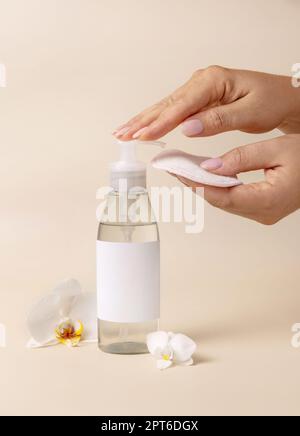 Una mano donna pompando liquido su un trucco rimuovere tampone da una bottiglia cosmetica vicino a fiori orchidee su beige primo piano, etichetta mockup. Produ di bellezza per la cura della pelle Foto Stock