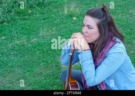 Bella ragazza bruna dai capelli lunghi vestita di blu seduta sull'erba guardando fuori dal profilo tenendo il suo ukulele Foto Stock