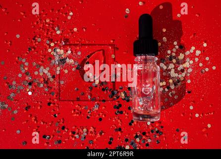 Flacone trasparente in vetro per siero o olio con pipetta. Sfondo rosso brillante con argento luccichio e coriandoli. Beffa. Concetto di cosmetica e. Foto Stock
