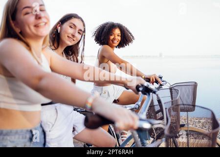 Adolescenti di diverse nazionalità e aspetto in bicicletta percorrono una strada cittadina. Le ragazze giovani e positive sorridono insieme. Stile di vita sano Foto Stock