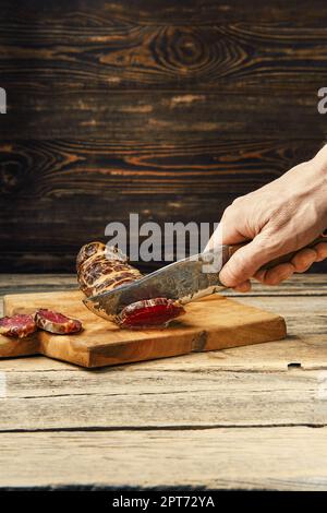 Mano con coltello che taglia carne di manzo affumicata su fette su tagliere di legno Foto Stock