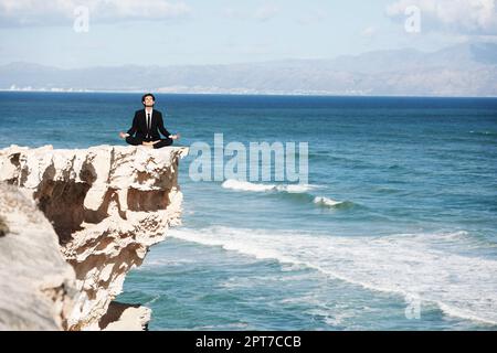 Trovare l'illuminazione in natura. Uomo d'affari seduto e meditato sulla cima di una scogliera sopra l'oceano Foto Stock