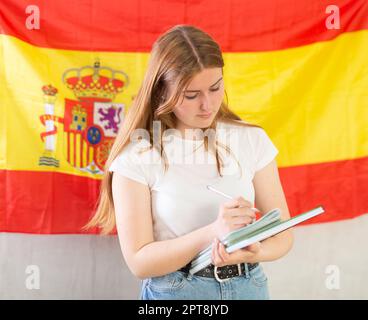 Ragazza giovane sorridente allegra, studente in abbigliamento casual scrivere in spirale contro di bandiera nazionale della Spagna Foto Stock