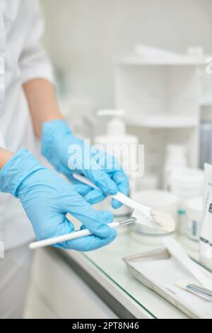 Primo piano di pennello bianco con crema viso di lusso in mani di estetista. Prodotti per la cura della pelle Foto Stock