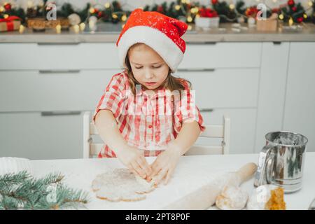 Bambina dai capelli scuri di 3 anni in berretto rosso di Natale e camicia a scacchi taglia i biscotti di pan di zenzero dalla pasta arrotolata in decoro bianco di Natale Foto Stock