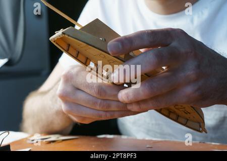 Mani di uomo incollando con colla i particolari del compensato per modello di nave, tenendo con le dita. Processo di costruzione di navi giocattolo, hobby, artigianato. Tavolo con variou Foto Stock