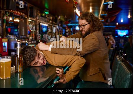 I tifosi di calcio combattono. Uomo aggressivo che tira il ragazzo sul bancone del bar. Amici conflitto in pub. Persone furiose Foto Stock