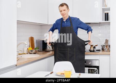 L'uomo adulto medio toglie il grembiule prima di mangiare la colazione che ha preparato da solo Foto Stock