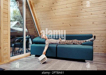 Uomo di mezza età che si appetta pacificamente sul divano nella sua casa di campagna. Ha lasciato il libro a terra. Foto Stock