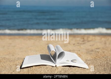 Libro aperto con pagine piegate a forma di cuore sulla spiaggia sabbiosa vicino al mare Foto Stock