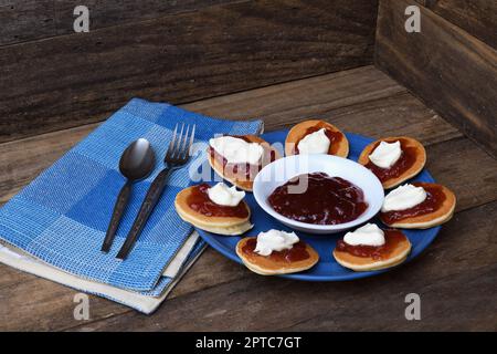 Pikelets/Pancakes/Battercakes con confettura di fragole e panna montata su un piatto blu con tovagliolo, cucchiaio e forchetta in un ambiente rustico in legno Foto Stock