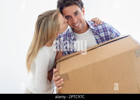 Dare una mano d'aiuto. Ritratto di un bel giovane sorridente e portatore di una scatola mentre la sua ragazza lo bacia sulla guancia Foto Stock