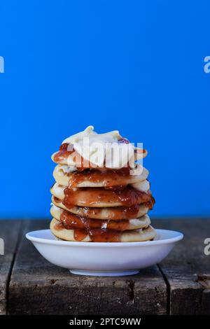 Una piccola ciotola bianca di pikelets/frittelle/tortine impilate l'una sull'altra con panna montata e marmellata di fragole che fuoriesce dai lati Foto Stock