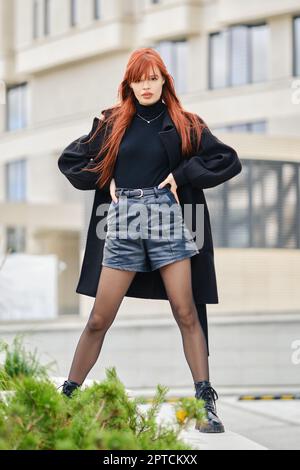 Giovane donna elegante con capelli rossi in cappotto nero, pullover e pantaloncini in pelle che si posano sulla strada Foto Stock