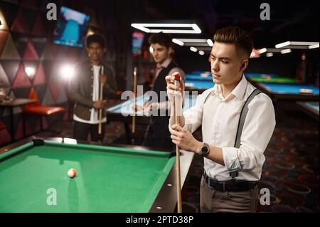 Giovani amici o studenti che giocano a biliardo, concentrarsi su un ragazzo elegante strofinando piscina cue Foto Stock