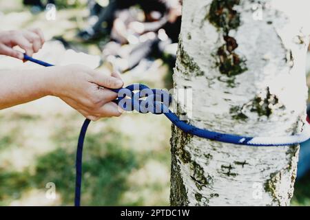 Le mani delle donne tirano una corda blu forte legata ad un albero di betulla. Installazione professionale di una zip line o di una cabinovia. Tempo di divertimento e relax. Weekend in famiglia Foto Stock