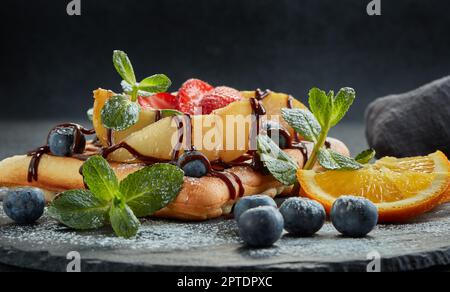 Deliziosi waffle con pera caramellata, frutti di bosco, menta, zucchero a velo e salsa dolce sul piano di servizio nero su sfondo grigio. Dessert. Serving foo Foto Stock