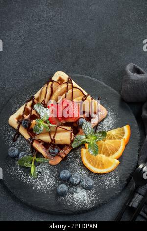 Deliziosi waffle con pera caramellata, frutti di bosco, menta, zucchero a velo e salsa dolce sul piano di servizio nero su sfondo grigio. Dessert. Serving foo Foto Stock