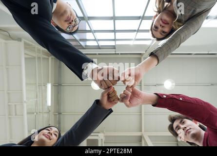 Concetto di team building. I colleghi si stanno in piedi in cerchio battendo il pugno insieme, celebrando il successo di affari in ufficio moderno con alto soffitto. Vie inferiore Foto Stock