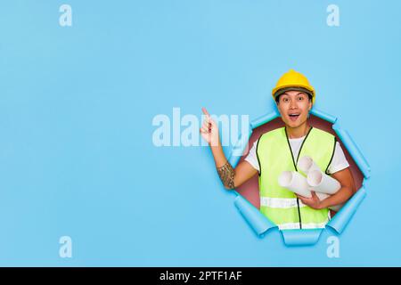 Giovane ingegnere asiatico isolato su sfondo blu indossando un elmetto giallo e tenendo in mano i rotoli di carta, sta puntando il dito sulla spac libera Foto Stock